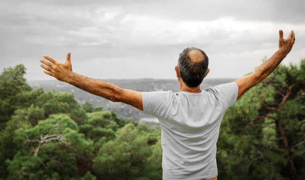 man standing above some trees with his back to the camera with arms extended, celebrating his practice of AA step 10